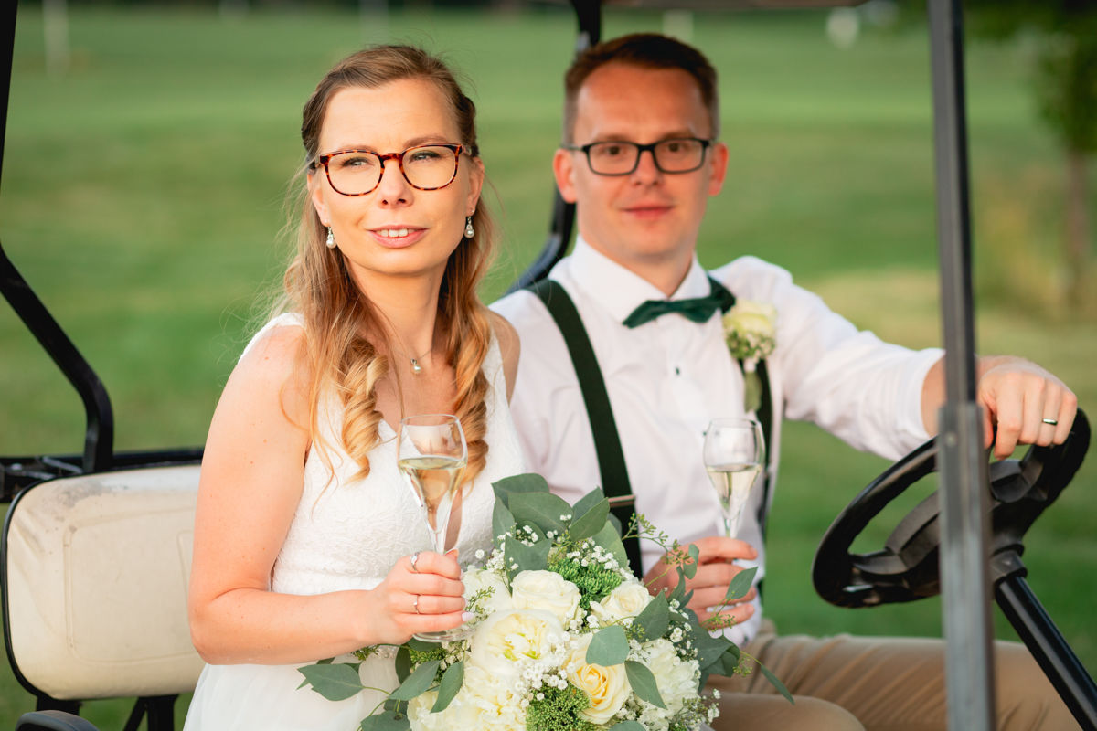 Nevěsta drží skleničku a jede se ženichem golfovým vozítkem